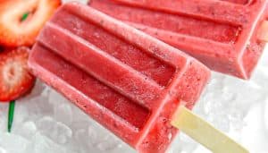 Two red strawberry popsicles on ice with sliced strawberries in the background.