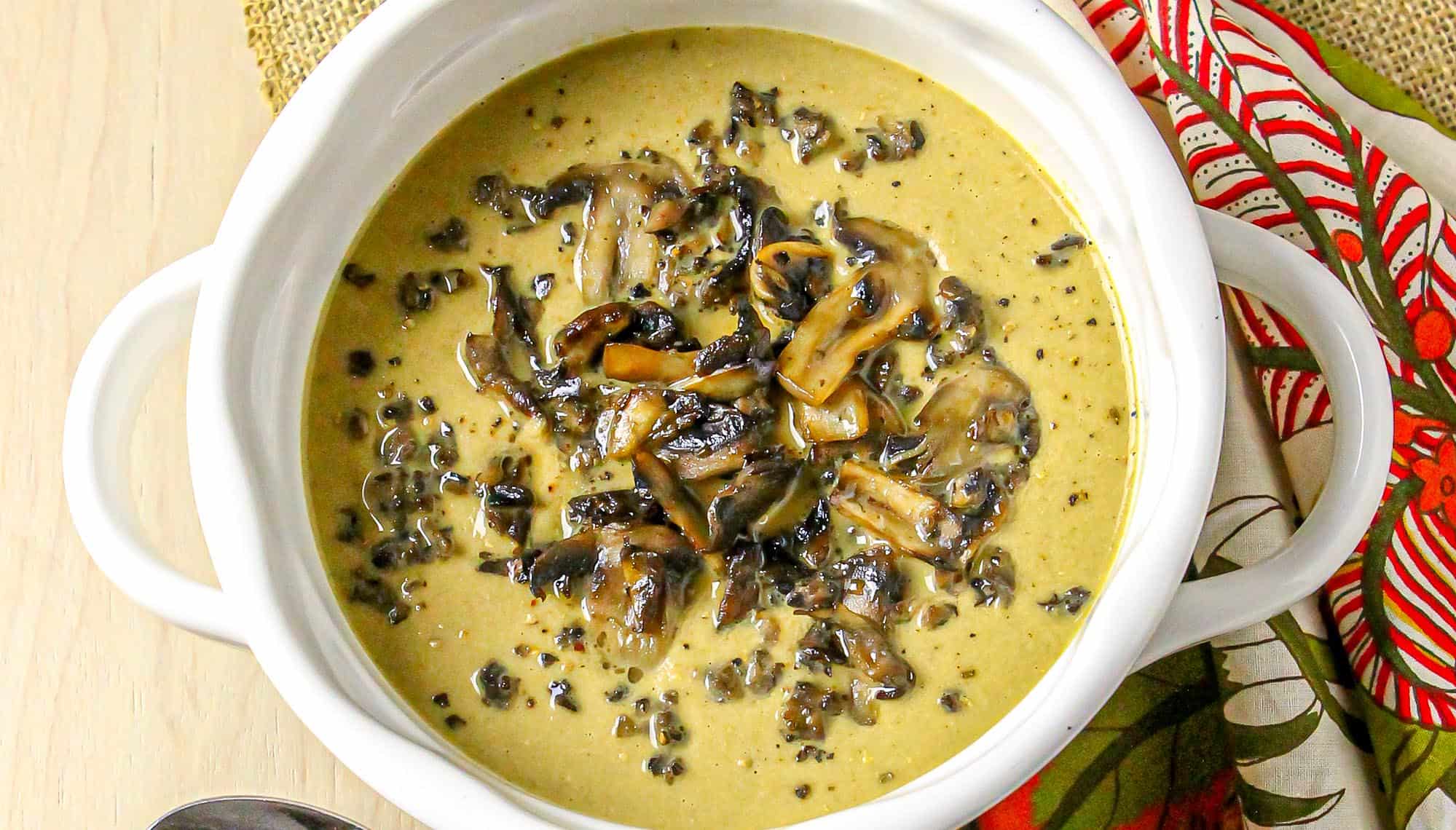 Creamy mushroom soup garnished with sautéed mushroom slices in a white bowl next to a patterned red and green cloth napkin.
