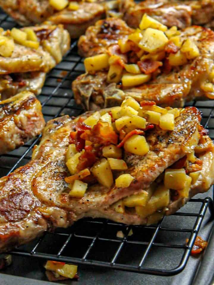 Pork chops topped with cooked diced apples and bacon on a black wire rack over a baking sheet.