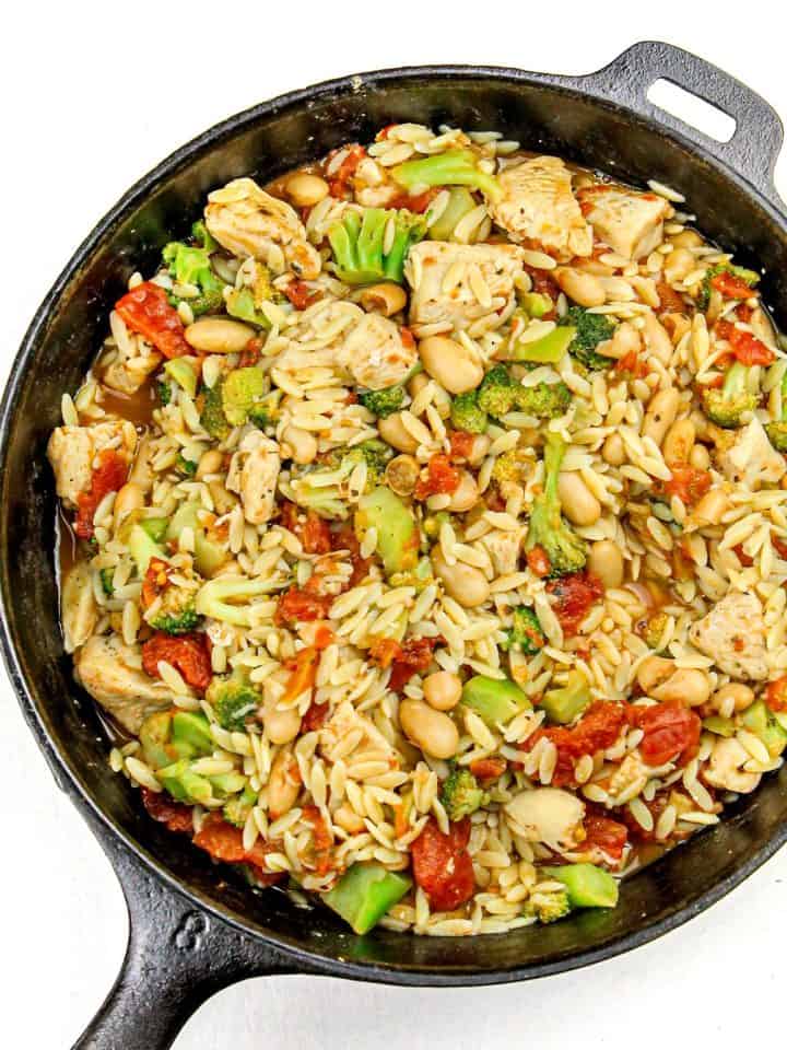 Cast iron skillet with cooked orzo, chicken pieces, broccoli florets, white beans, and diced tomatoes.