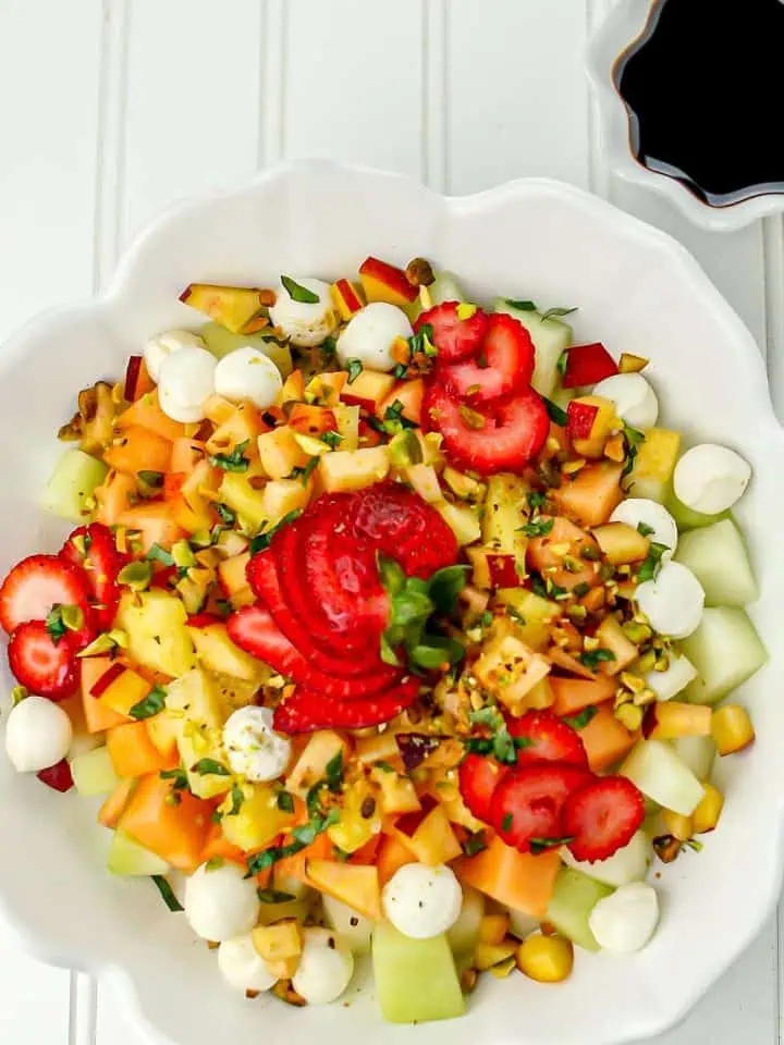 Fresh fruit salad with strawberries, cantaloupe, pineapple, melon, mozzarella balls, pistachios, and herbs in a white scalloped bowl with a small dish of dark dressing
