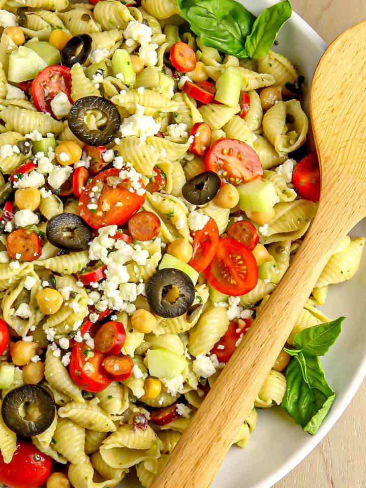 Pasta salad with cherry tomatoes, black olives, chickpeas, cucumber, feta cheese, basil leaves, and wooden spoon in a white bowl.