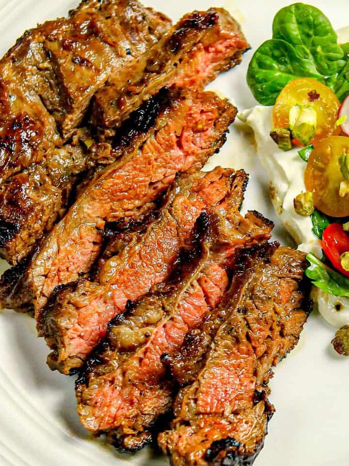 Sliced medium-rare grilled steak with a side salad of spinach, cherry tomatoes, radishes, pistachios, and whipped ricotta on a white plate.