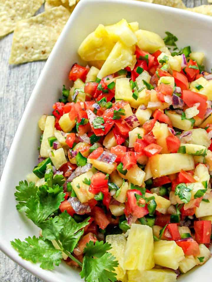 Bowl of pineapple salsa with diced tomatoes, red onion, jalapeño, cilantro, and tortilla chips in the background on wooden surface.