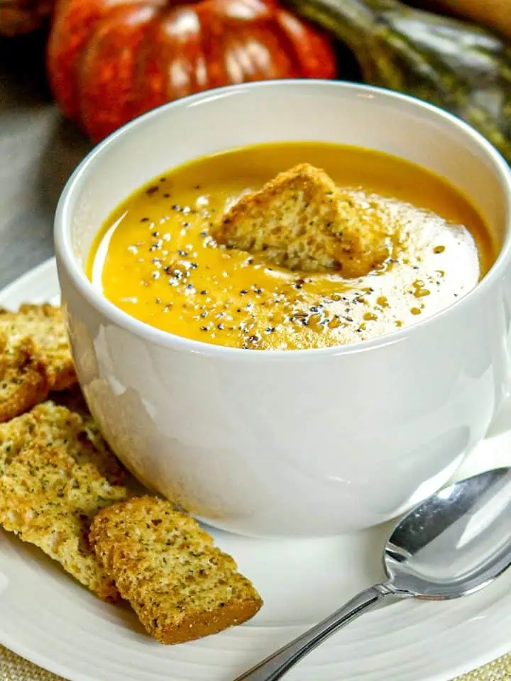 Creamy butternut squash soup in a white mug topped with a crouton, served with herb crackers on a plate next to a spoon, with gourds in the background.
