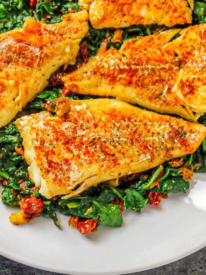 Pan-seared seasoned fish fillets served on a bed of sautéed spinach with sun-dried tomatoes on a white plate.