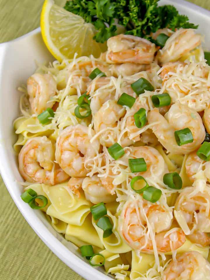 Shrimp pasta with creamy sauce, shredded cheese, chopped green onions, lemon wedge, and parsley garnish in white bowl on green placemat.