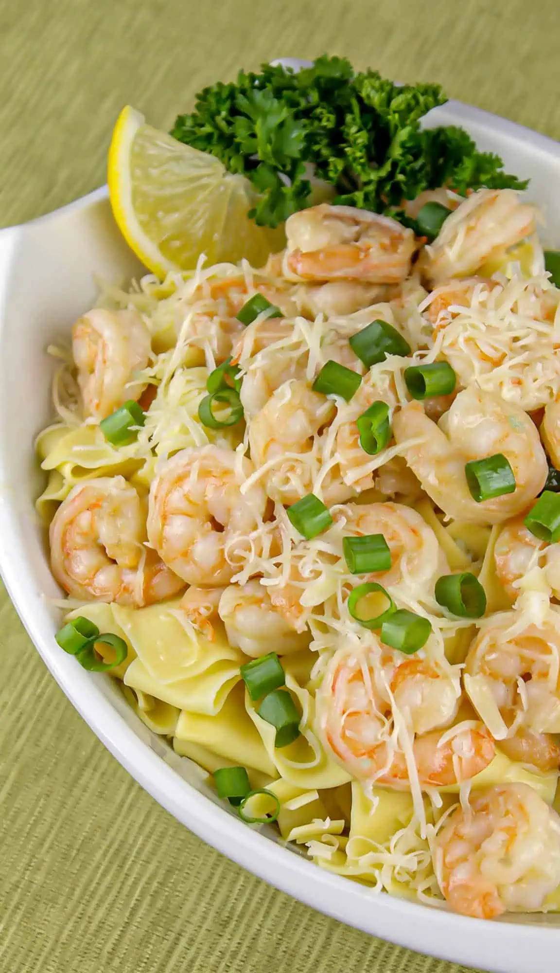 Shrimp pasta with creamy sauce, shredded cheese, chopped green onions, lemon wedge, and parsley garnish in white bowl on green placemat.