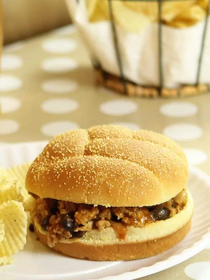 Sloppy joe sandwich on a sandwich bun with wavy potato chips on a white plate, with a basket of chips in the background.