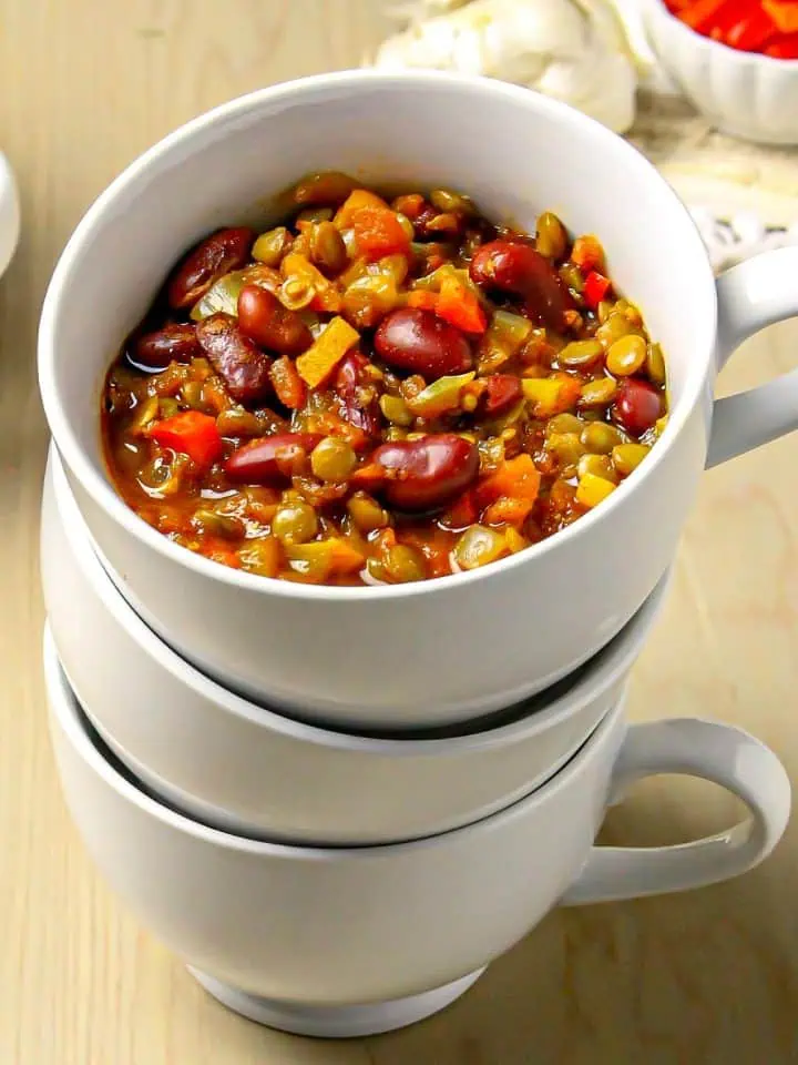Lentil and kidney bean chili with diced vegetables served in a white cup stacked on two empty white cups on a wooden table.