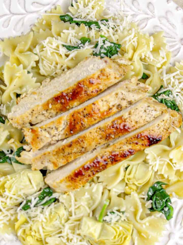 Grilled chicken breast slices over bowtie pasta with spinach, artichoke hearts, and shredded cheese on a decorative white plate.