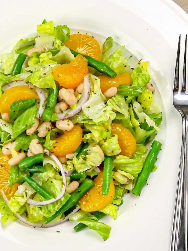 Salad with green beans, mandarin orange slices, white beans, red onion, and lettuce on a white plate with a fork beside it.