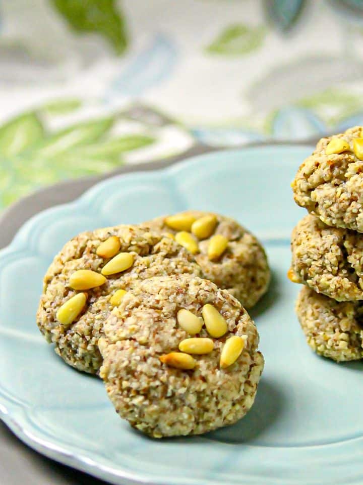 Close-up of round almond cookies topped with pine nuts on a blue plate with a floral-patterned tablecloth in the background.
