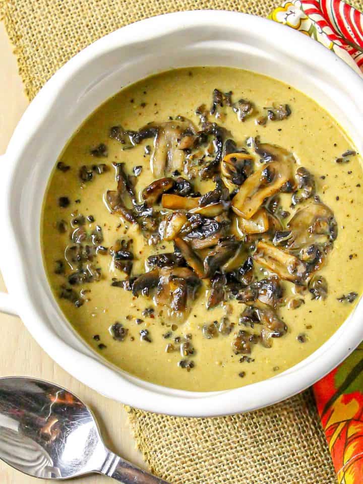 Creamy mushroom soup in white bowl with sautéed mushrooms on top, placed on burlap cloth and colorful napkin, with metal spoon beside.
