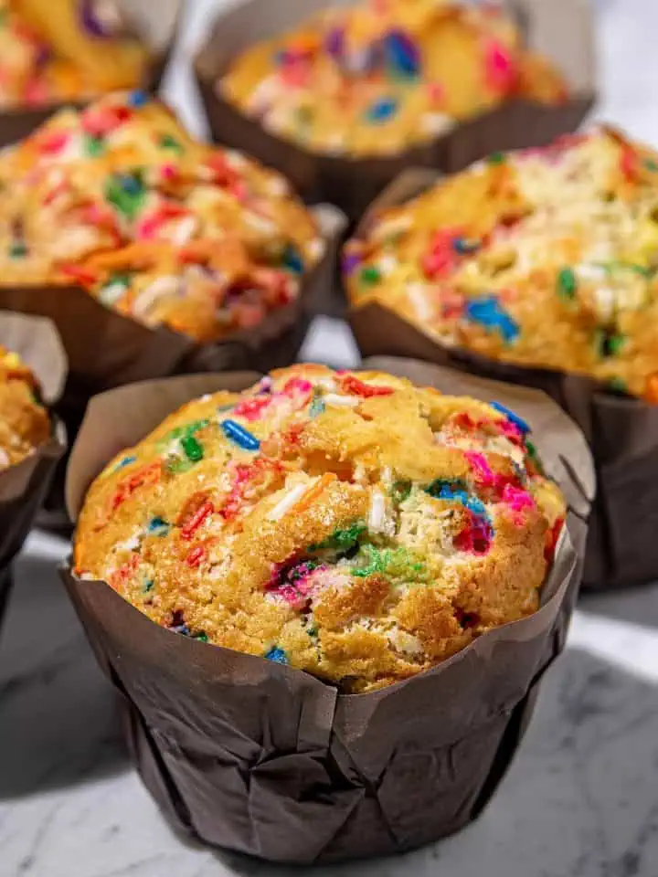Colorful confetti muffins in dark brown paper liners on a marble surface, with focus on the front muffin.