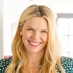 Smiling woman with long blonde hair wearing a green floral top in a bright indoor setting.