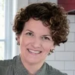 Woman with short curly hair smiling, wearing a greenish-gray top, with a tiled wall and window in the background.