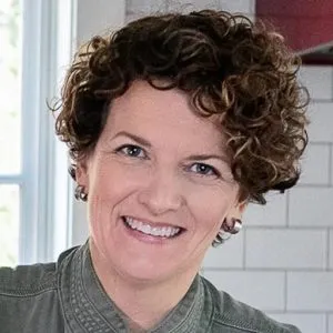 Woman with short curly hair smiling, wearing a greenish-gray top, with a tiled wall and window in the background.