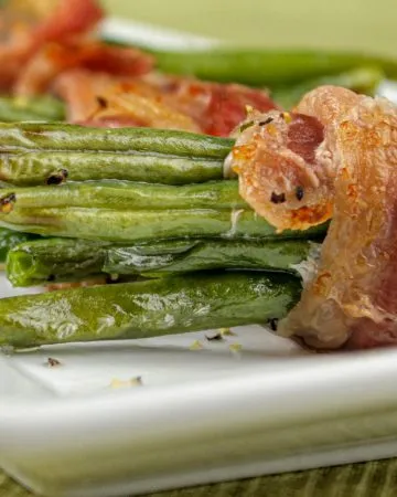 Green bean bundles wrapped in crispy bacon on a white rectangular plate with a green textured background.