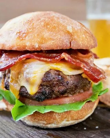 Burger with bacon, melted cheese, lettuce, and tomato on a toasted bun, served with potato chips and a pickle wedge on a wooden board.