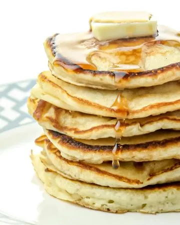 Stack of six golden-brown pancakes topped with a pat of melting butter and dripping maple syrup on a white plate with gray geometric rim.