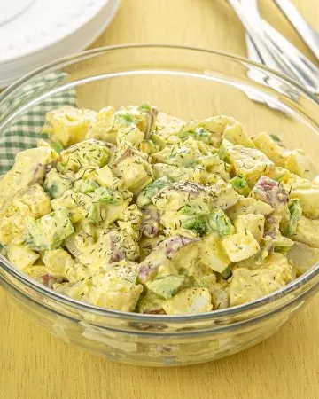 Bowl of creamy potato salad with herbs, celery, and red onion on a wooden table with green checkered cloth and stacked plates with forks.