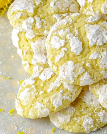 Stack of yellow lemon crinkle cookies dusted with powdered sugar on a white surface with lemon zest sprinkled around.
