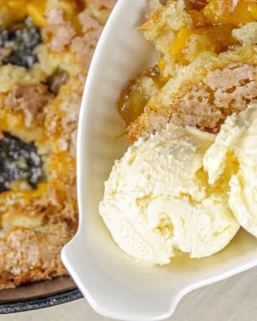 Peach cobbler in a cast iron skillet alongside a serving of cobbler with two scoops of vanilla ice cream on a white dish.