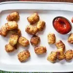 Crispy tater tots arranged on a white tray with a small dish of ketchup and some ketchup drops on a wooden surface.