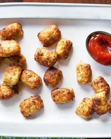 Crispy tater tots arranged on a white tray with a small dish of ketchup and some ketchup drops on a wooden surface.