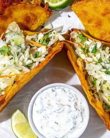 Crispy tacos filled with cabbage slaw and a side of creamy herb sauce with lime wedges on a metal tray.