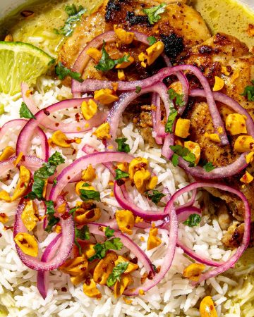 Grilled chicken thigh with white rice, sliced lime, pickled red onions, chopped nuts, and herbs in a green sauce in a white bowl.