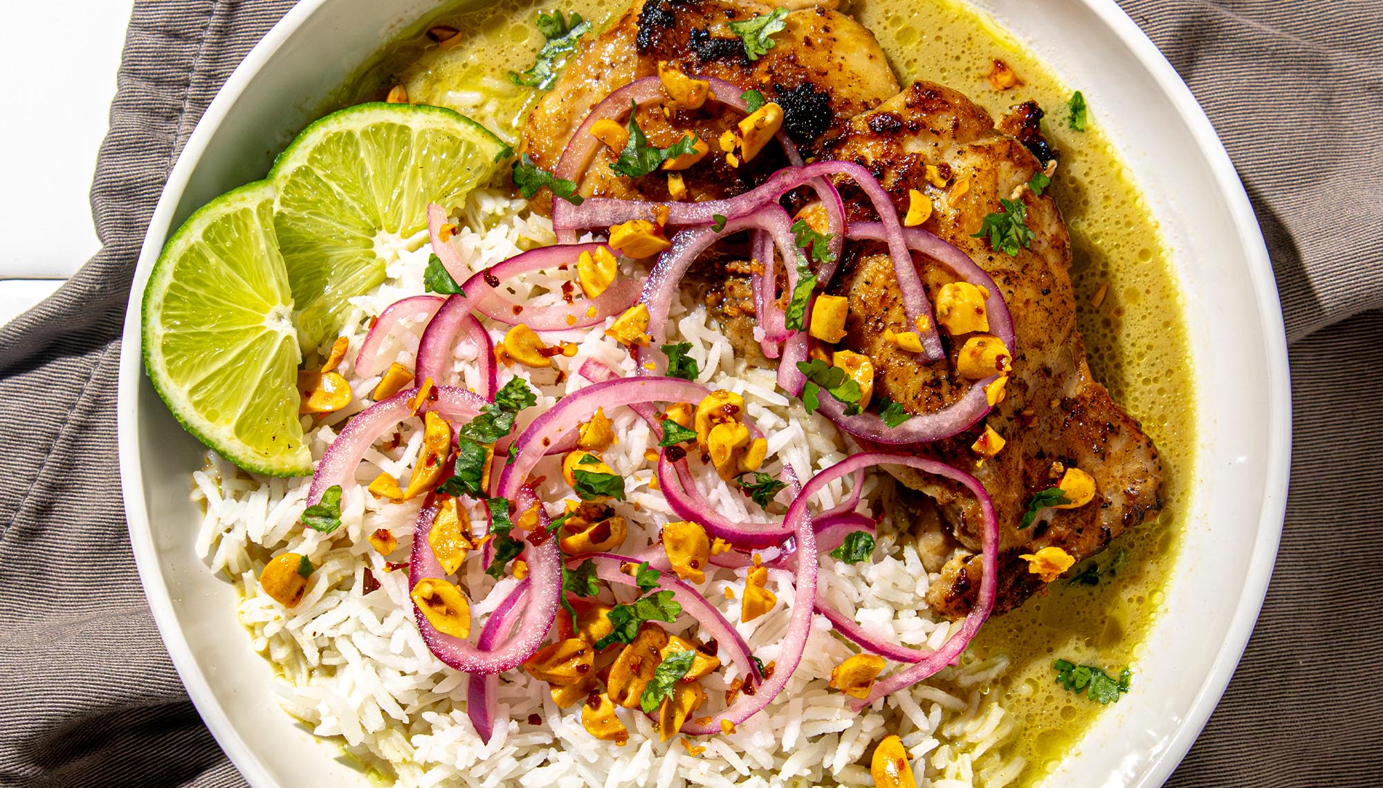 Grilled chicken thigh with white rice, sliced lime, pickled red onions, chopped nuts, and herbs in a green sauce in a white bowl.