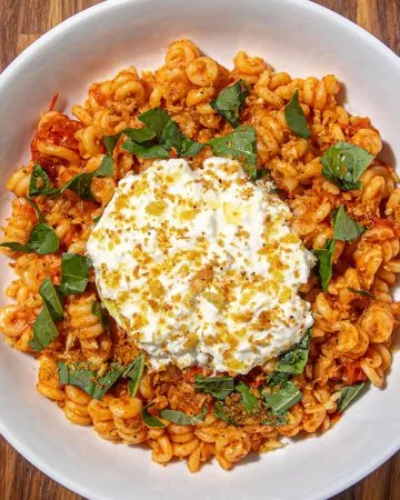 Fusilli corti pasta with tomato sauce, fresh basil, Burrata cheese, and breadcrumb topping in a white bowl on a wooden surface.