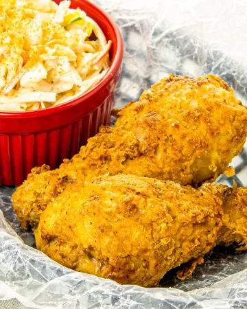 Two pieces of crispy fried chicken drumsticks with a side of creamy coleslaw in a red ramekin on wax paper.