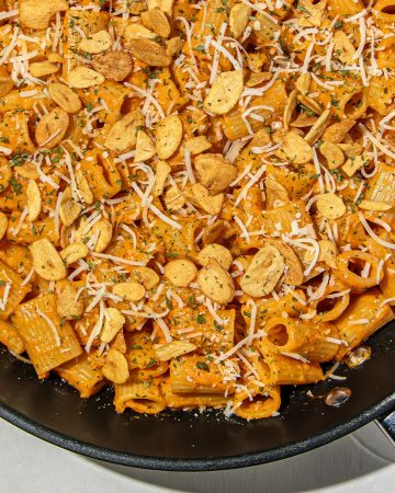 Rigatoni pasta in creamy gochujang sauce topped with shredded cheese, dried herbs, and crispy garlic chips in a black skillet.