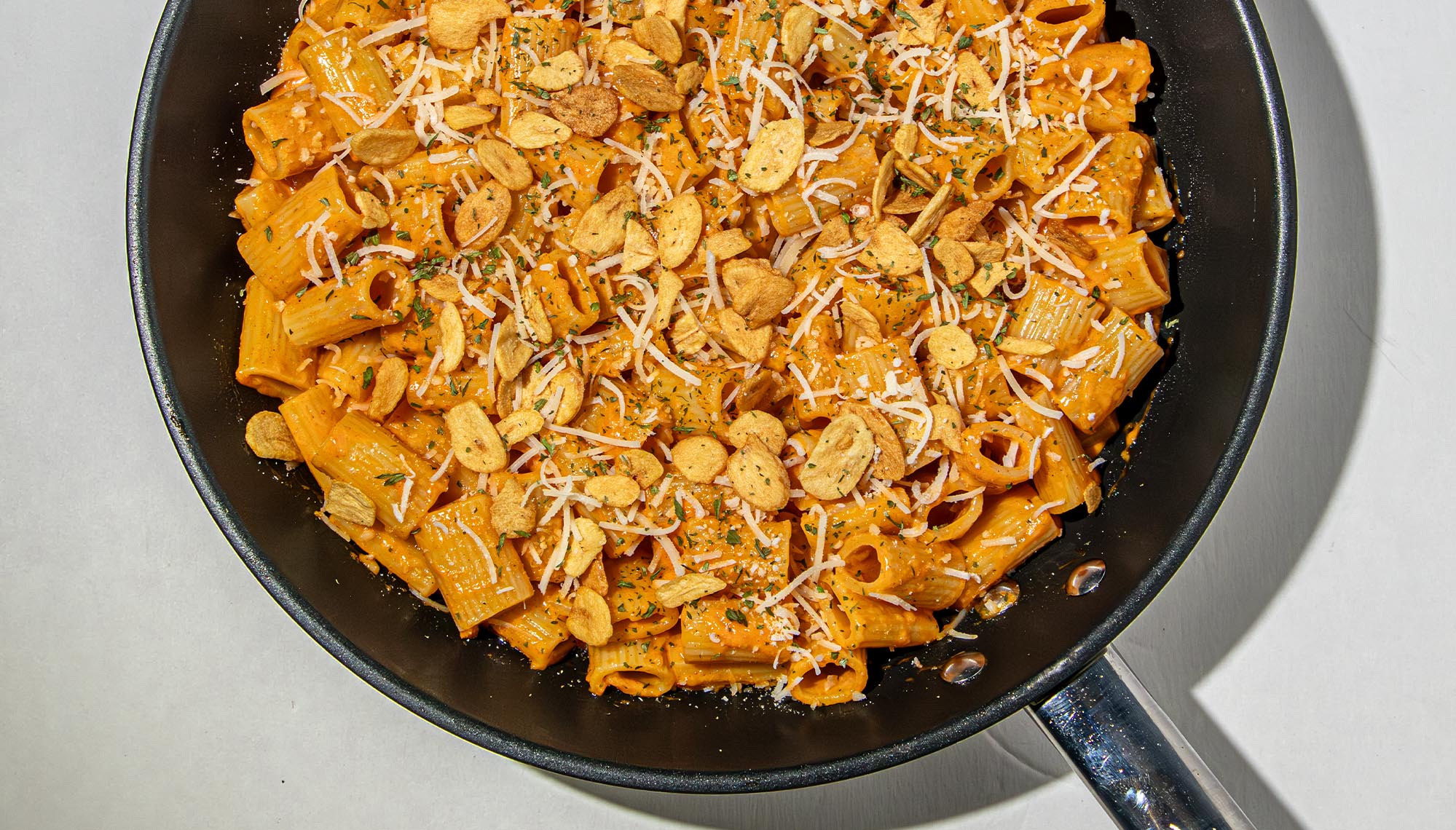 Rigatoni pasta in creamy gochujang sauce topped with shredded cheese, dried herbs, and crispy garlic chips in a black skillet.