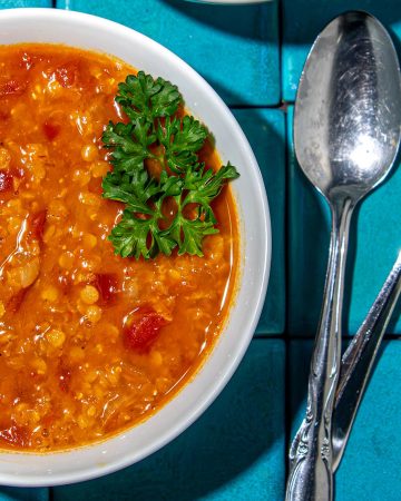 Tomato lentil soup garnished with parsley in white bowl, next to two silver spoons on blue tiled surface.