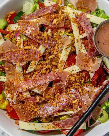 Salad with mixed greens, cucumber, tomato, sliced meats, cheese strips, crispy topping, dressing on the side, and fork in a white bowl on marble surface.