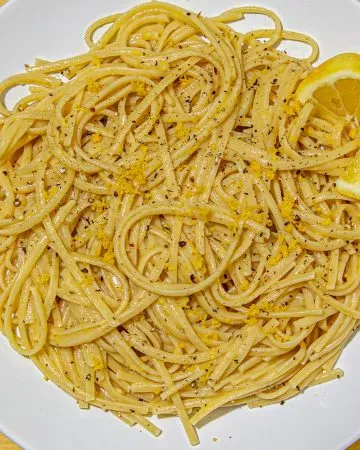 Linguine pasta tossed with lemon zest and black pepper, garnished with a lemon wedge on a white plate over a yellow wooden surface.