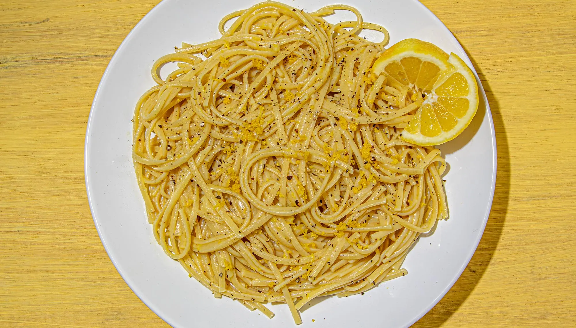 Linguine pasta tossed with lemon zest and black pepper, garnished with a lemon wedge on a white plate over a yellow wooden surface.