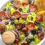 Salad with pepperoni, black olives, cherry tomatoes, green bell peppers, red onions, banana peppers, shredded cheese, dressing, and two garlic bread slices.