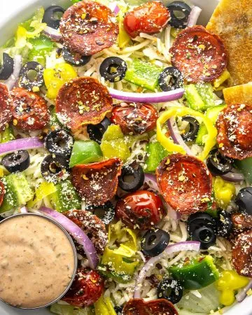 Salad with pepperoni, black olives, cherry tomatoes, green bell peppers, red onions, banana peppers, shredded cheese, dressing, and two garlic bread slices.
