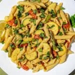Penne pasta with asparagus, zucchini, cherry tomatoes, peas, and fresh basil on a white plate with a green wooden background.