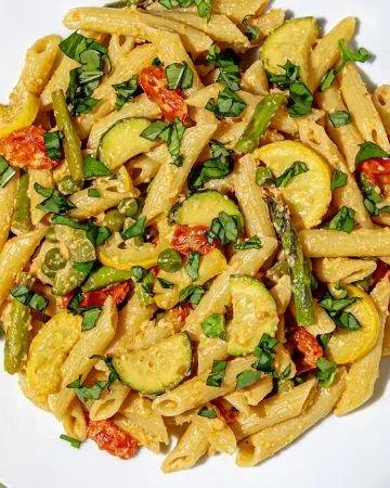 Penne pasta with asparagus, zucchini, cherry tomatoes, peas, and fresh basil on a white plate with a green wooden background.