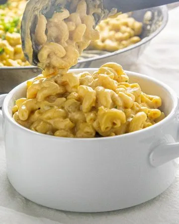 Creamy macaroni and cheese being served with tongs into a white bowl on a striped cloth.