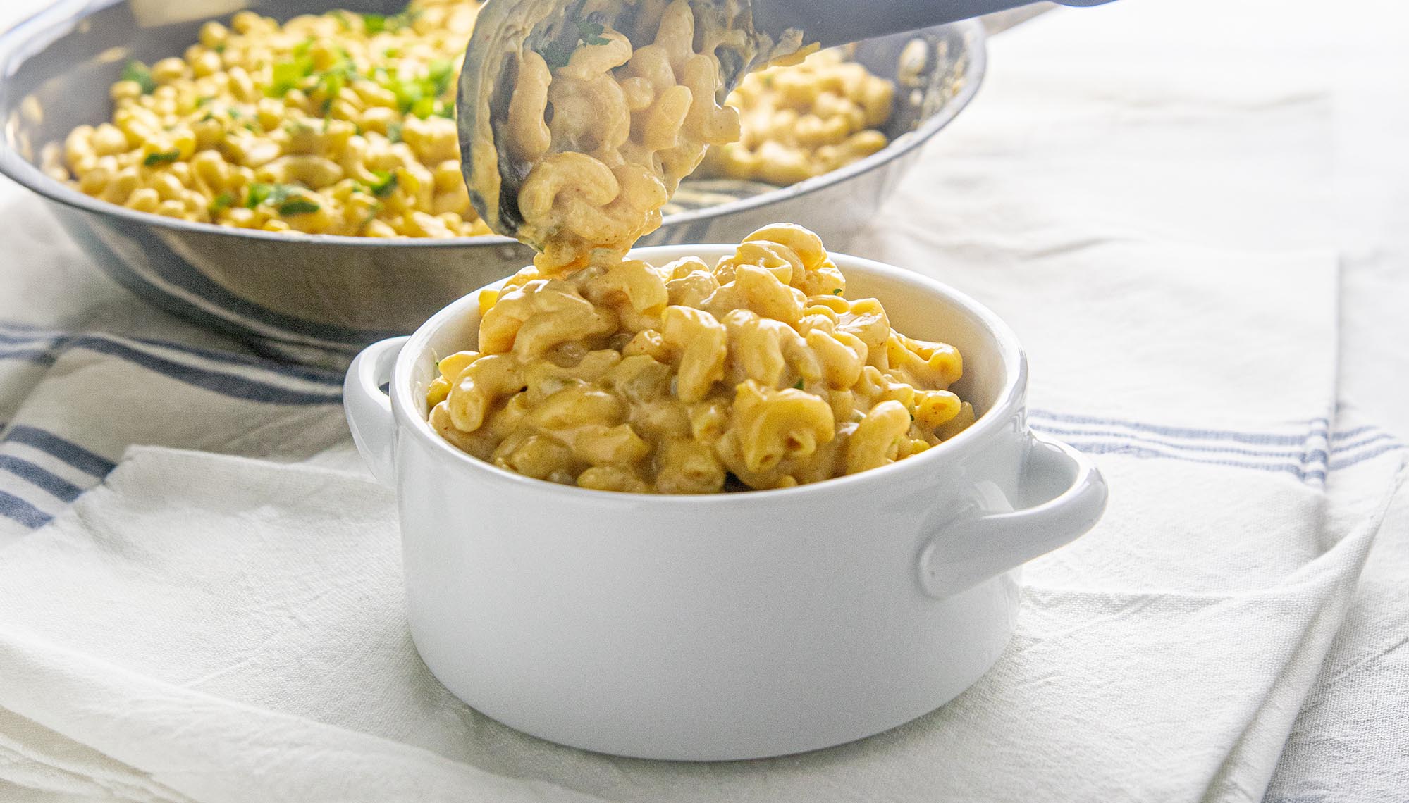 Creamy macaroni and cheese being served with tongs into a white bowl on a striped cloth.