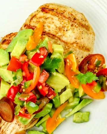 Grilled chicken breast topped with fresh salsa of diced tomatoes, avocado, bell peppers, and cilantro on a white plate with a patterned cloth.