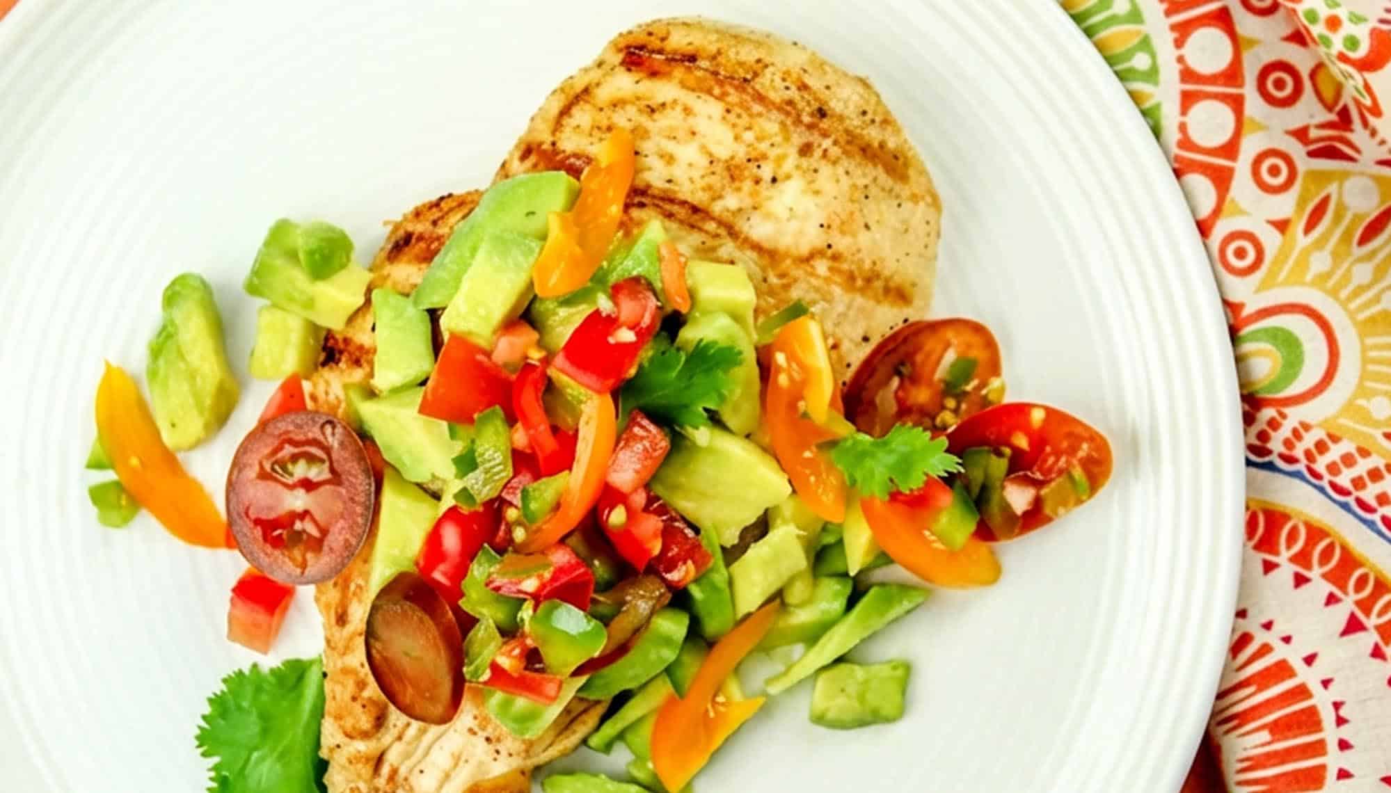 Grilled chicken breast topped with fresh salsa of diced tomatoes, avocado, bell peppers, and cilantro on a white plate with a patterned cloth.