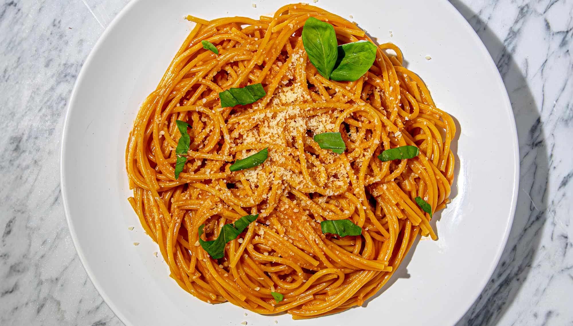 Spaghetti with tomato sauce, sprinkled with grated cheese and fresh basil leaves on a white plate over marble surface.
